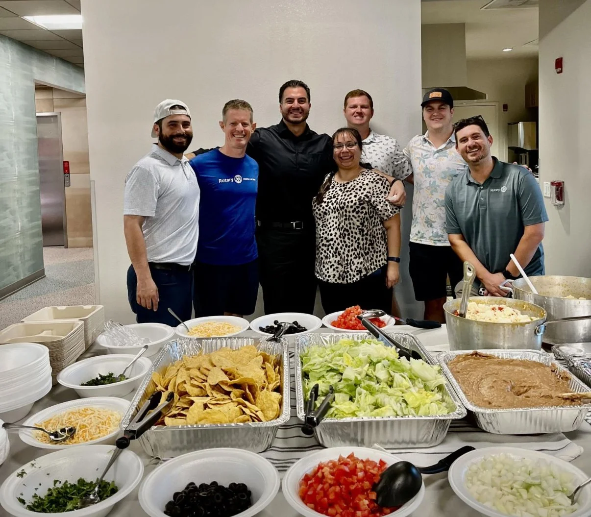 Group of seven people smiling behind a table of food, including chips, lettuce, tomato, black olives, shredded cheese, and various toppings, at a buffet-style gathering. At a Ronald McDonald House Charities event.