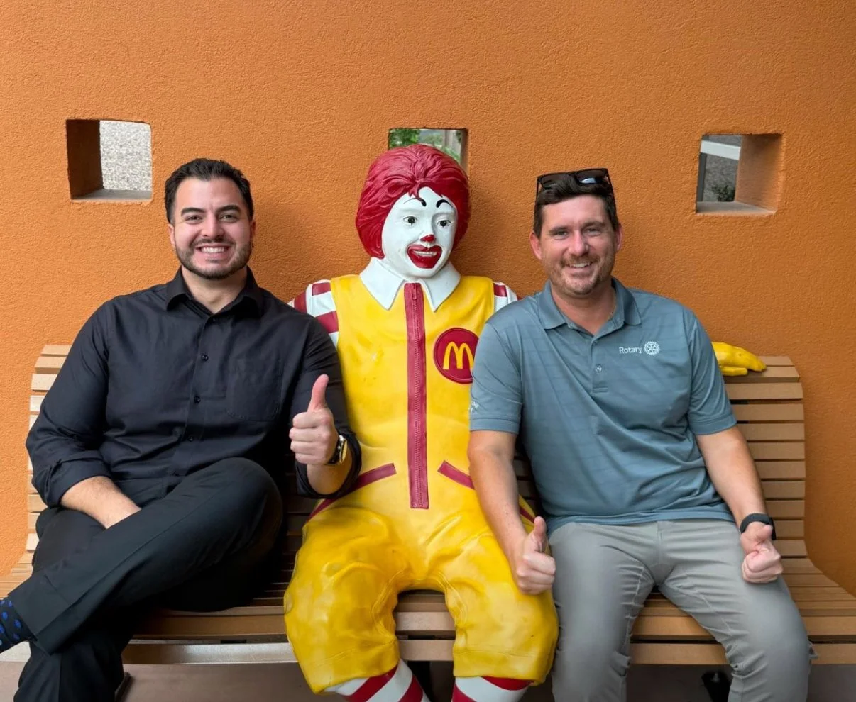 Two men sitting on a wooden bench next to a Ronald McDonald statue against an orange wall with small square windows. The man on the left is wearing a black shirt and giving a thumbs-up; the man on the right is wearing a gray polo shirt and glasses, a