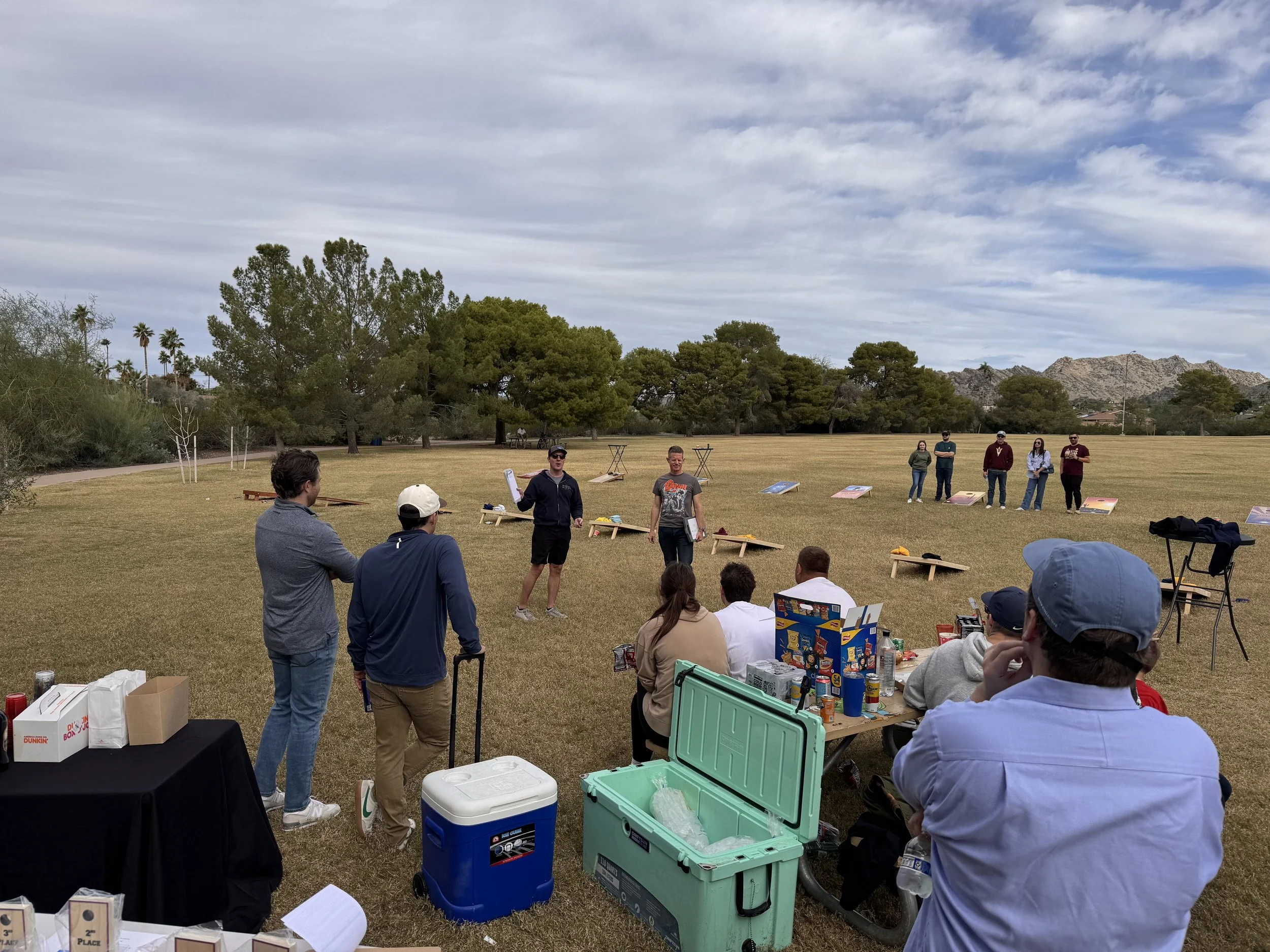 People gathered outdoors on a grassy field with a clear sky, participating in a cornhole game event. Some individuals are standing and watching, while others are sitting at a table with drinks and supplies.