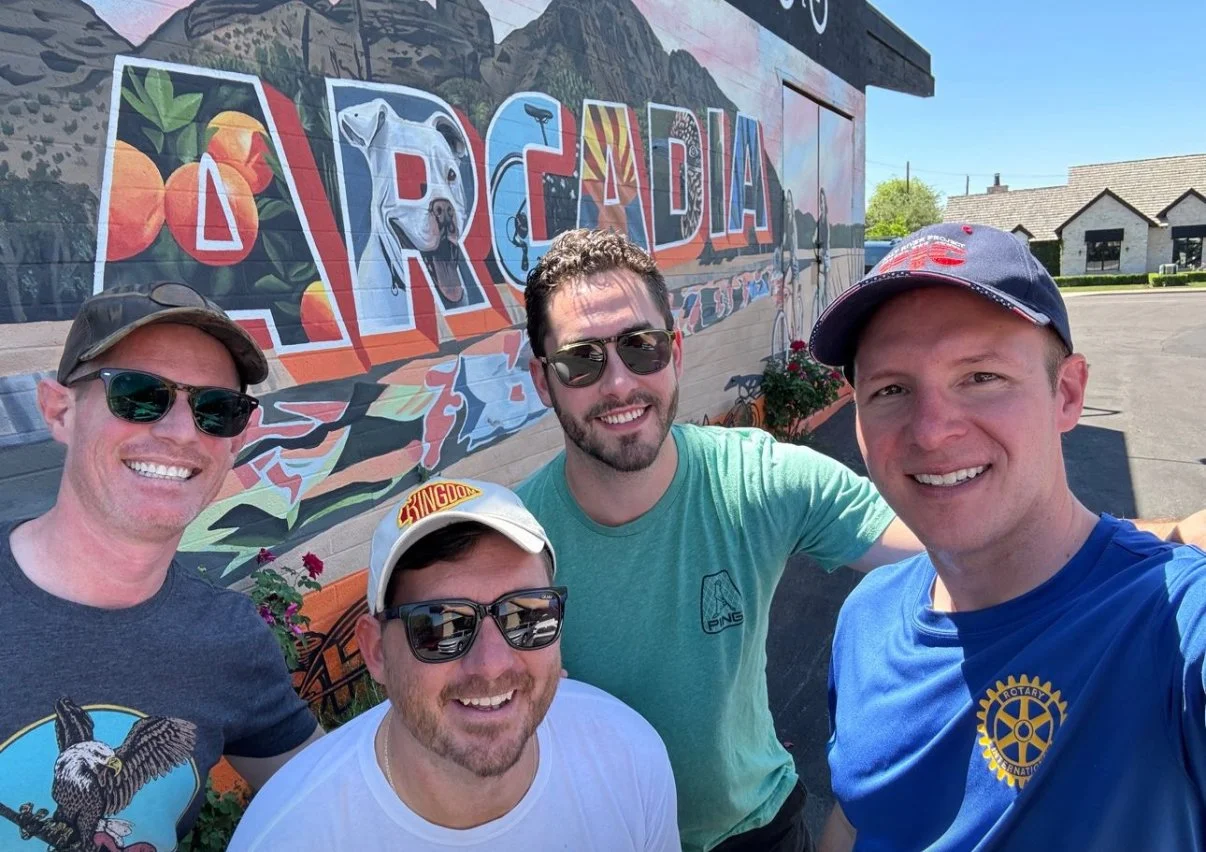 Four men smiling for a selfie in front of a colorful mural with the word 'ARCADIA' painted on it. The mural features vibrant artwork including a dog, oranges, and abstract patterns. The men are wearing casual clothing and sunglasses, and there's a bu