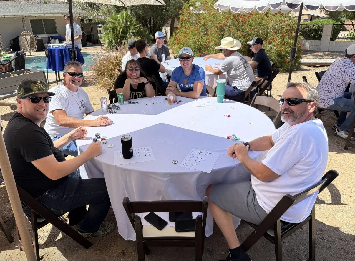 A group of people sitting around a round table outdoors, enjoying a social gathering. Some are playing poker or a card game, with chips and papers on the table. The setting is sunny with shade provided by umbrellas, and there are plants and a swimmin