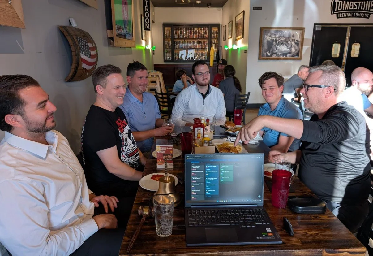 A group of six men sitting around a wooden table at a restaurant or bar, engaged in conversation. Tombstone Brewery Weekly Happy Hour Meeting Thursday nights at 6PM-7PM.