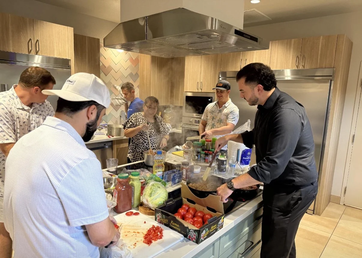 Group of people cooking and preparing food together in a modern kitchen, with ingredients and utensils on the counter. At a Ronald McDonald House Charities event.