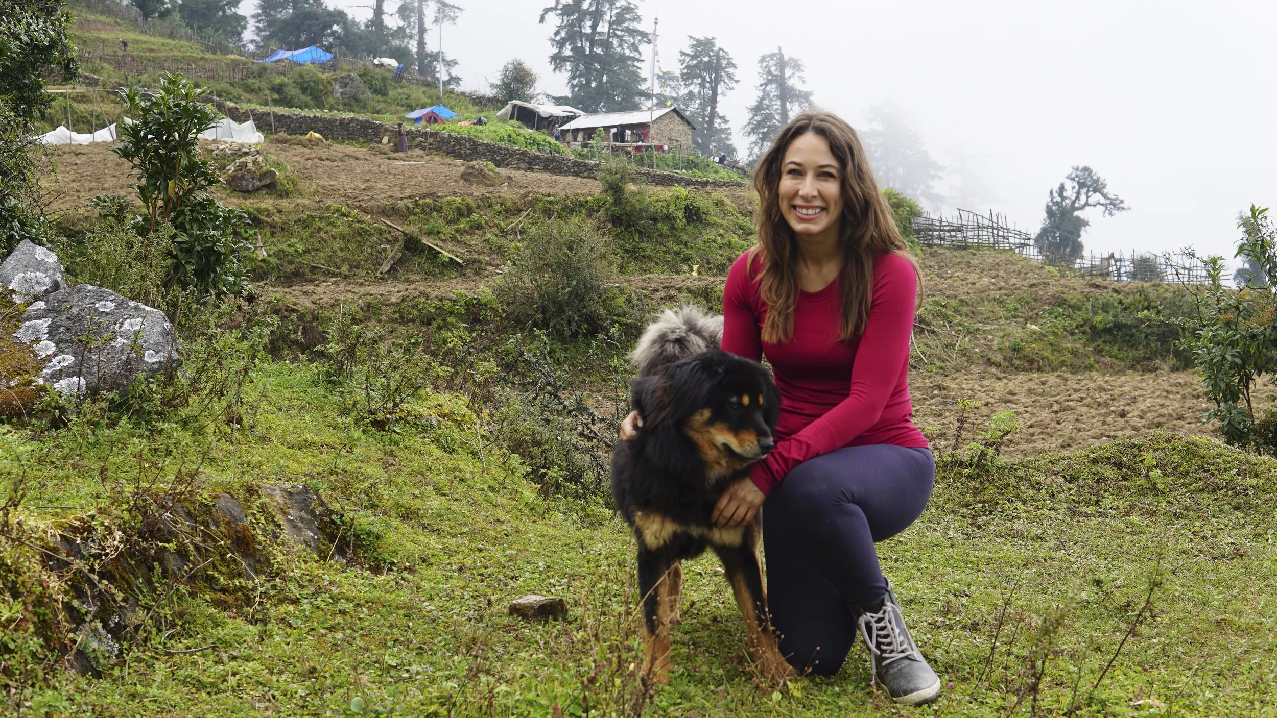 Temple Dog Rescue founder Lesley Mapstone kneels beside a black and brown dog in Nepal's green, mist-covered hills.