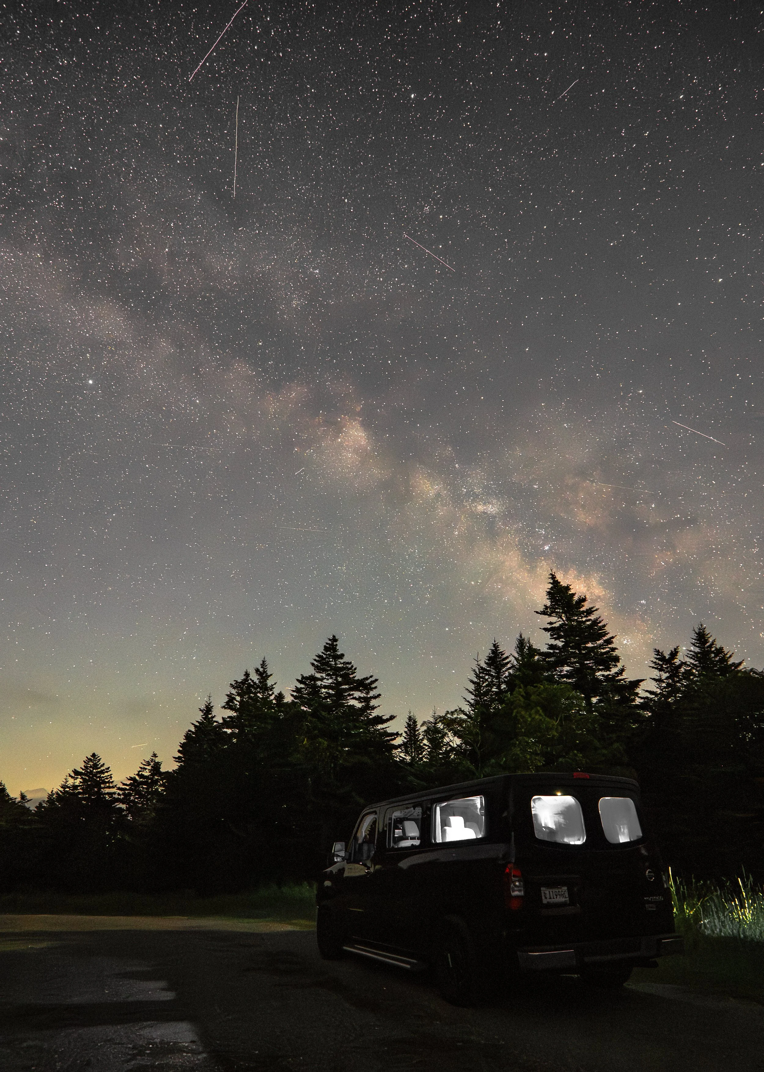 Night sky filled with stars and the Milky Way galaxy above a forested area, with a black camper van parked on a gravel surface in the foreground.