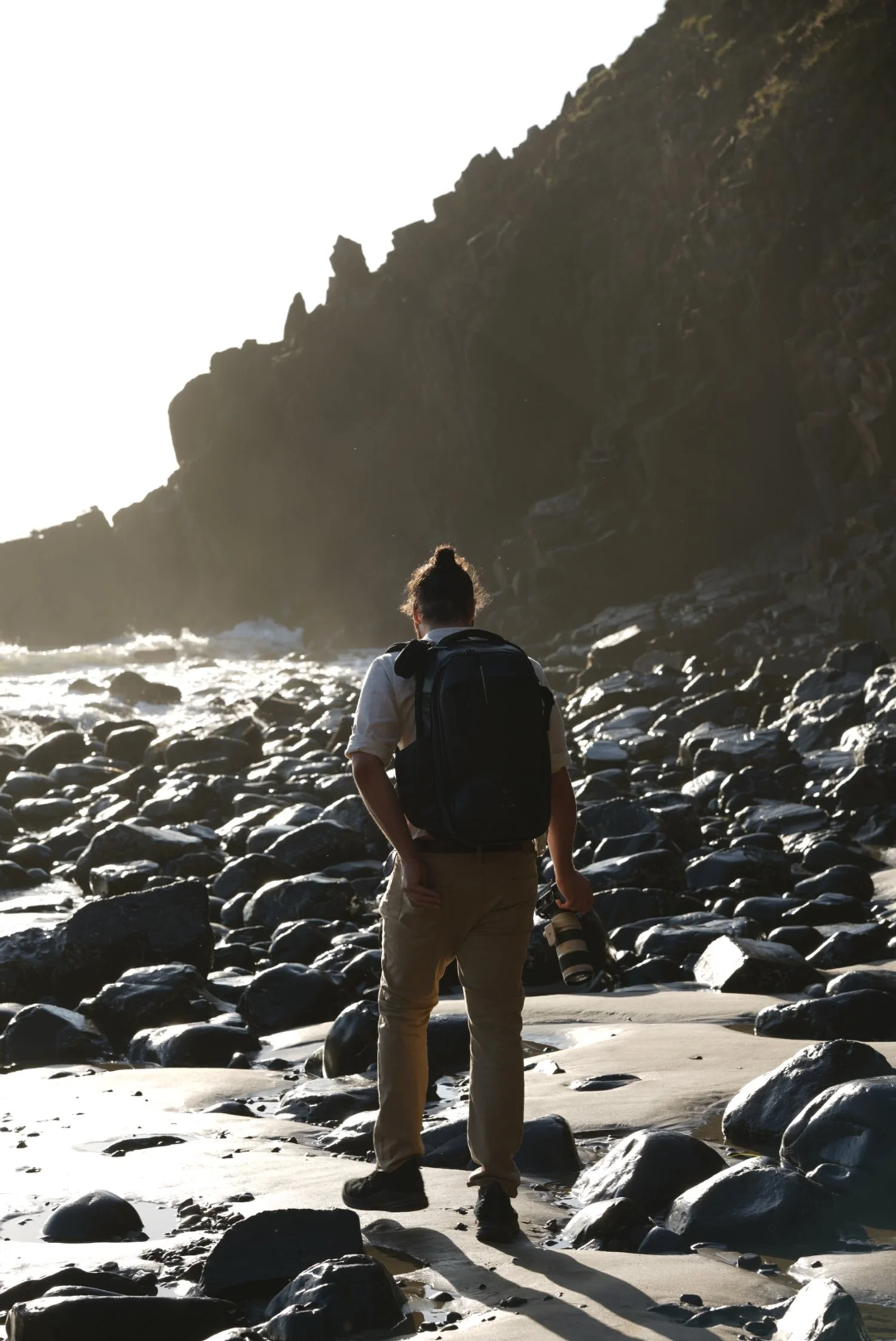 A person with a backpack and a camera standing on a rocky beach facing the ocean at sunset.