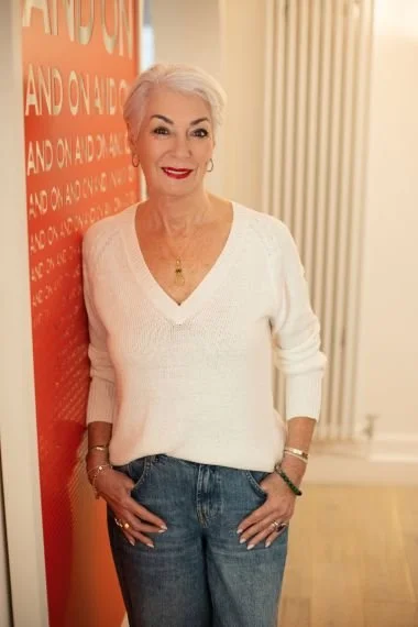 An elderly woman with white hair, wearing a white V-neck sweater and blue jeans, standing indoors next to a red wall with text, smiling at the camera.