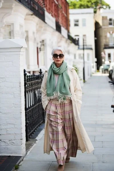 An elderly woman walking on a city sidewalk, wearing sunglasses, a beige trench coat, a green scarf, and plaid pants, with buildings and parked cars in the background.