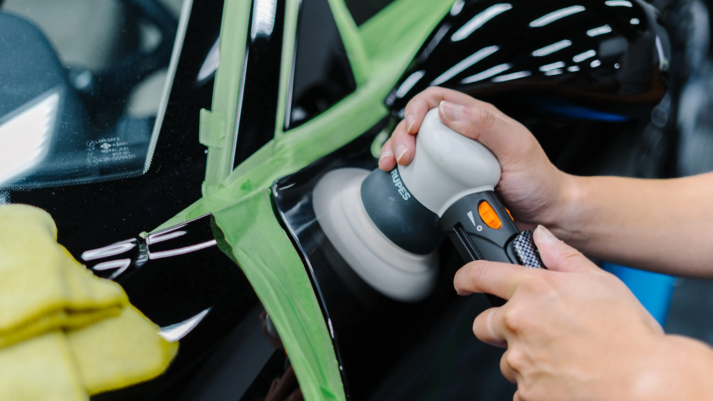 A person using a power buffer to polish a black car's windshield, with masking tape around the edges to protect surrounding areas, and a yellow microfiber cloth nearby.