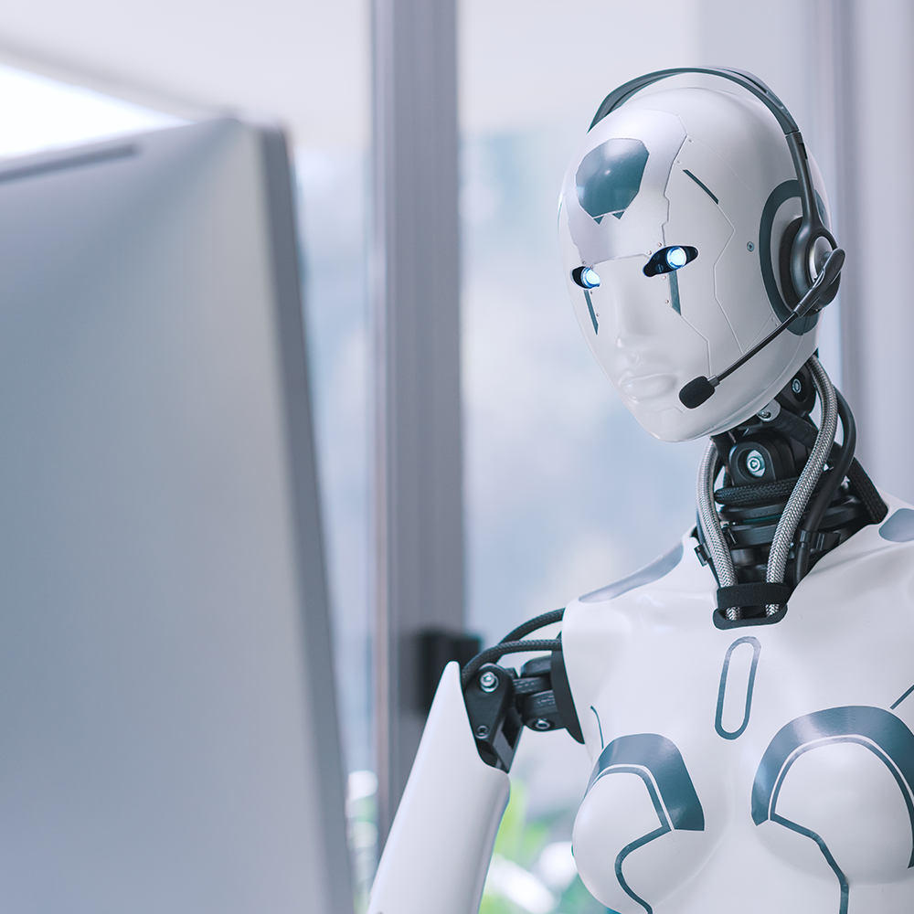 A humanoid robot wearing a headset, sitting in front of a computer monitor.