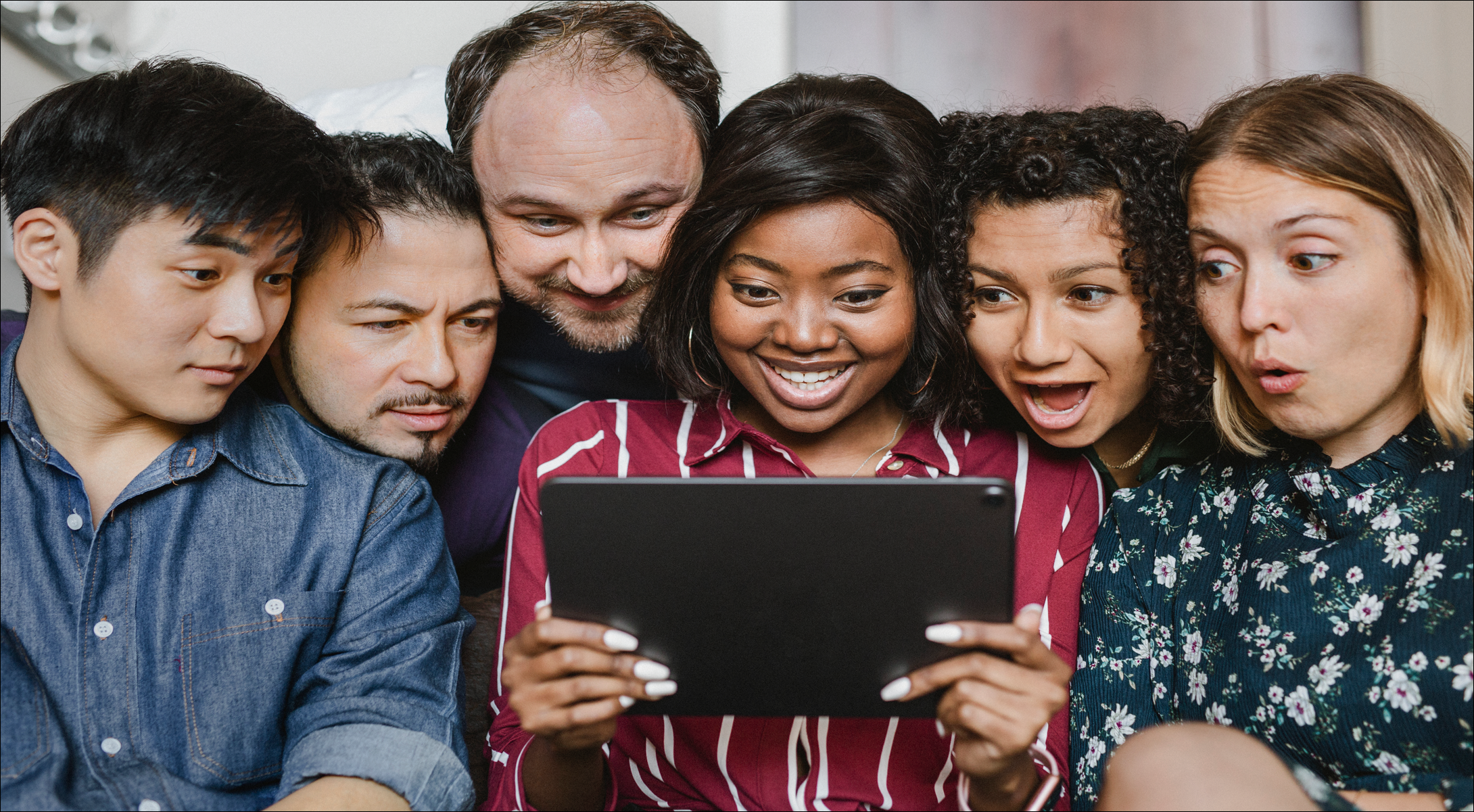 Group of six diverse young people gathered closely, looking at a tablet held by a woman in the middle, smiling and surprised.