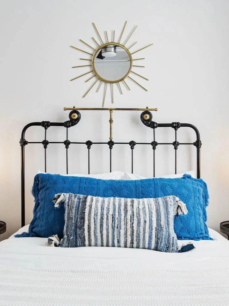 A bedroom with a black metal headboard, blue and striped pillows, a white bedspread, a mirrored sunburst wall decor, and a round side table.