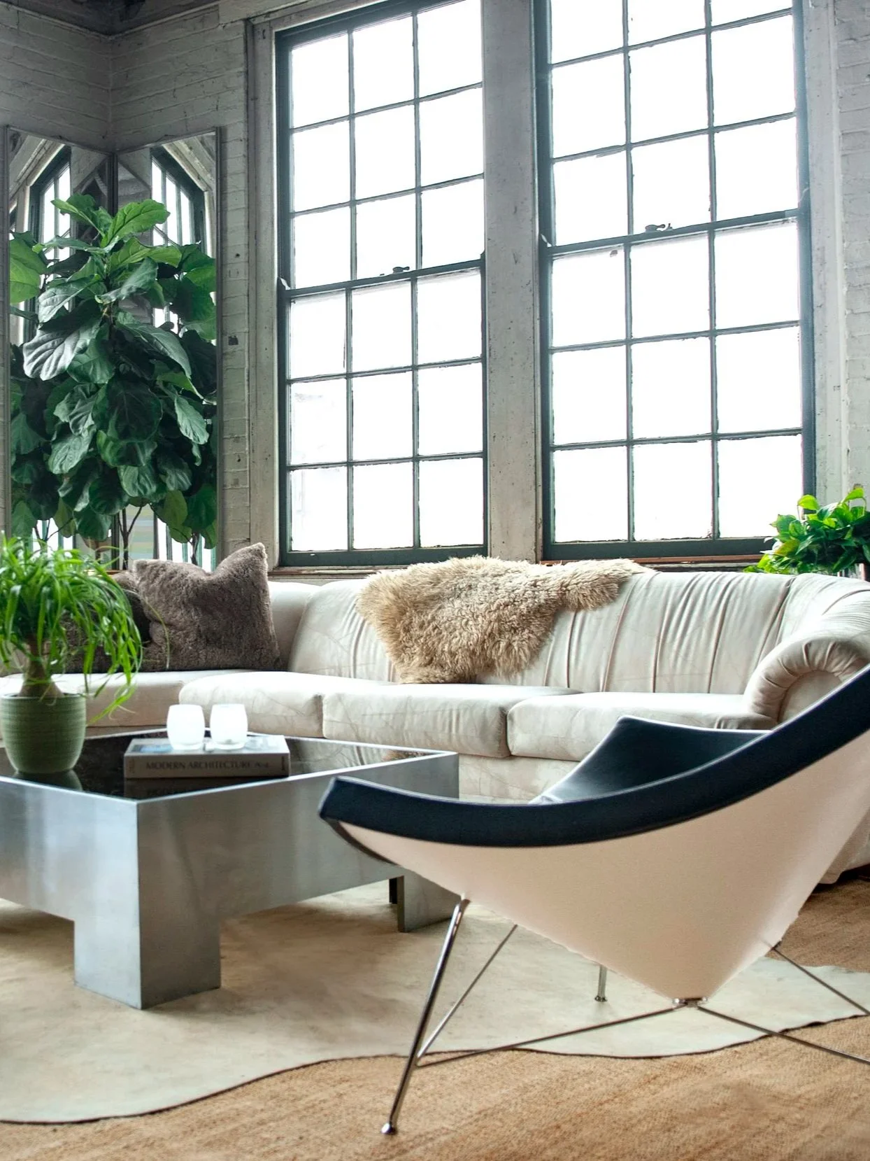 Modern living room with large industrial-style windows, a white sofa with textured pillows and a throw blanket, a black and white lounge chair, a glass coffee table with a plant and books, and greenery in the corner.
