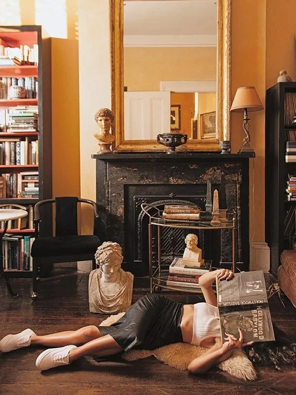 A person lying on the floor in front of a fireplace, reading a book, with bust sculptures and a large mirror hanging above the fire