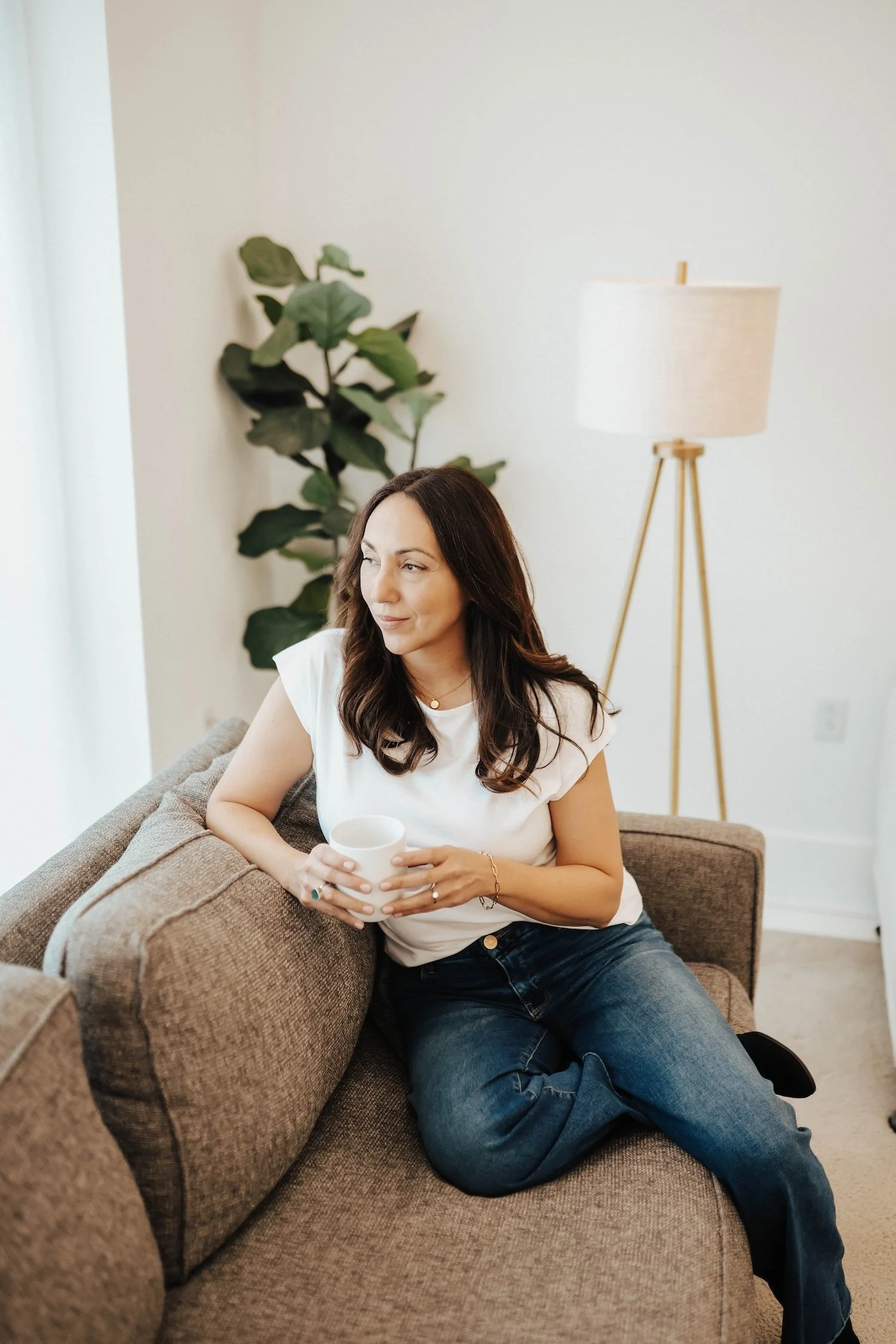 A woman with dark brown hair and light skin, wearing a white shirt and blue jeans, sitting on a beige couch holding a white mug, in a well-lit living room with a green plant and a tall white floor lamp in the background.