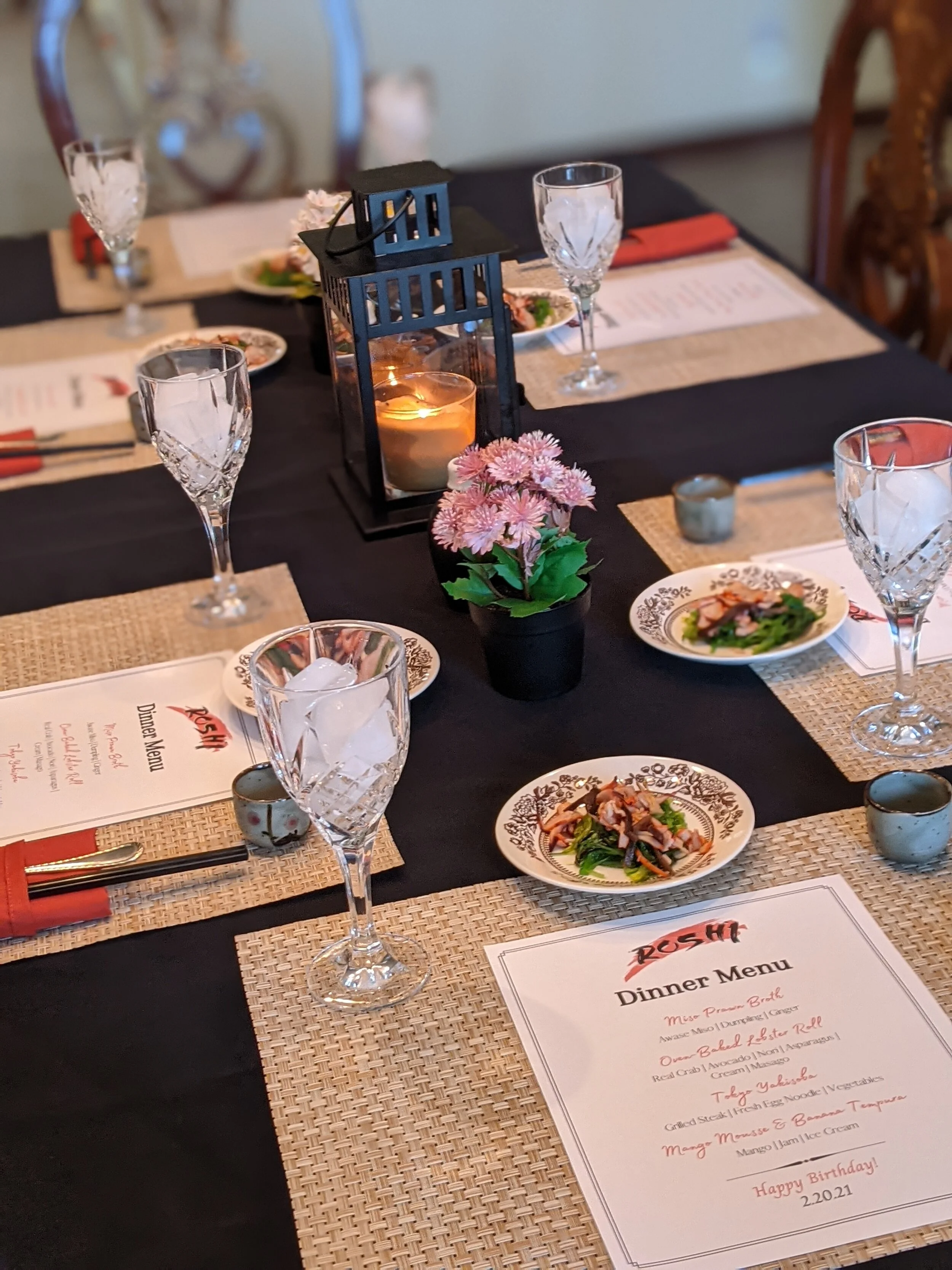 A dining table set for a meal, with plates of food, wine glasses, a candle inside a lantern, a small potted flower, and a dinner menu.