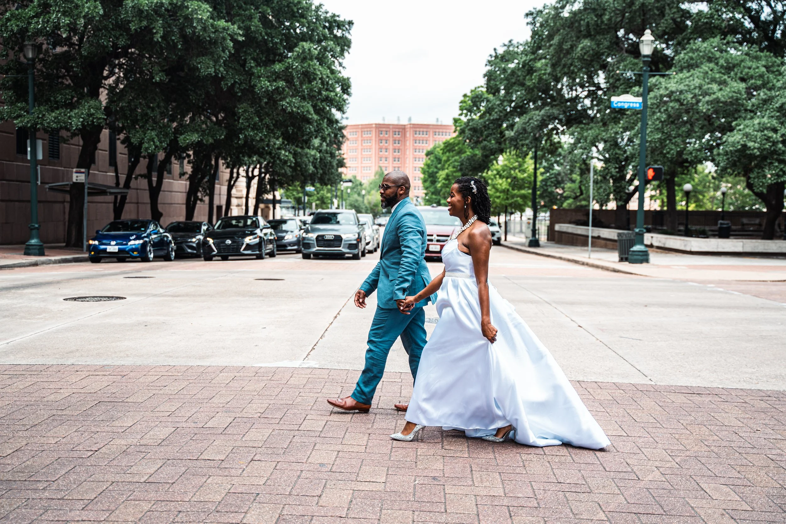 A bride and groom, holding hands and walking across a city street, with the bride in a white wedding gown and the groom in a blue suit.