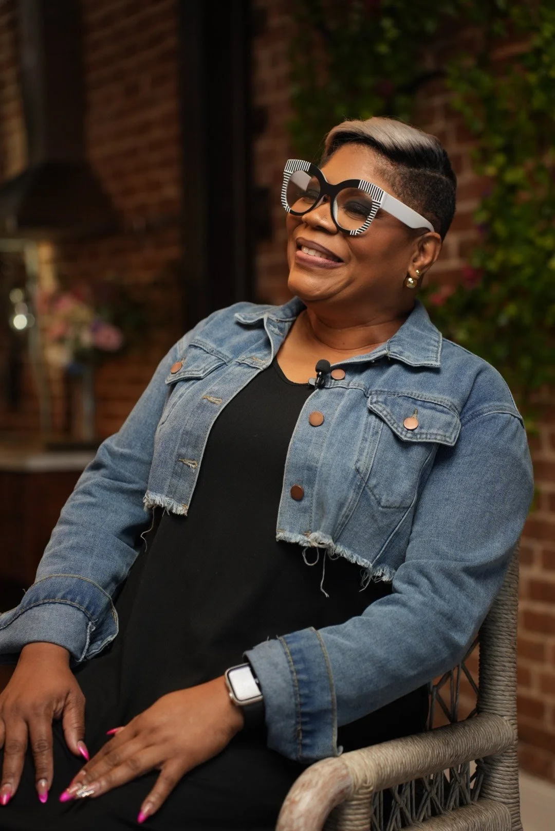 A woman with short hair and glasses smiling, wearing a denim jacket over a black top, sitting outside, with a brick wall and greenery in the background.