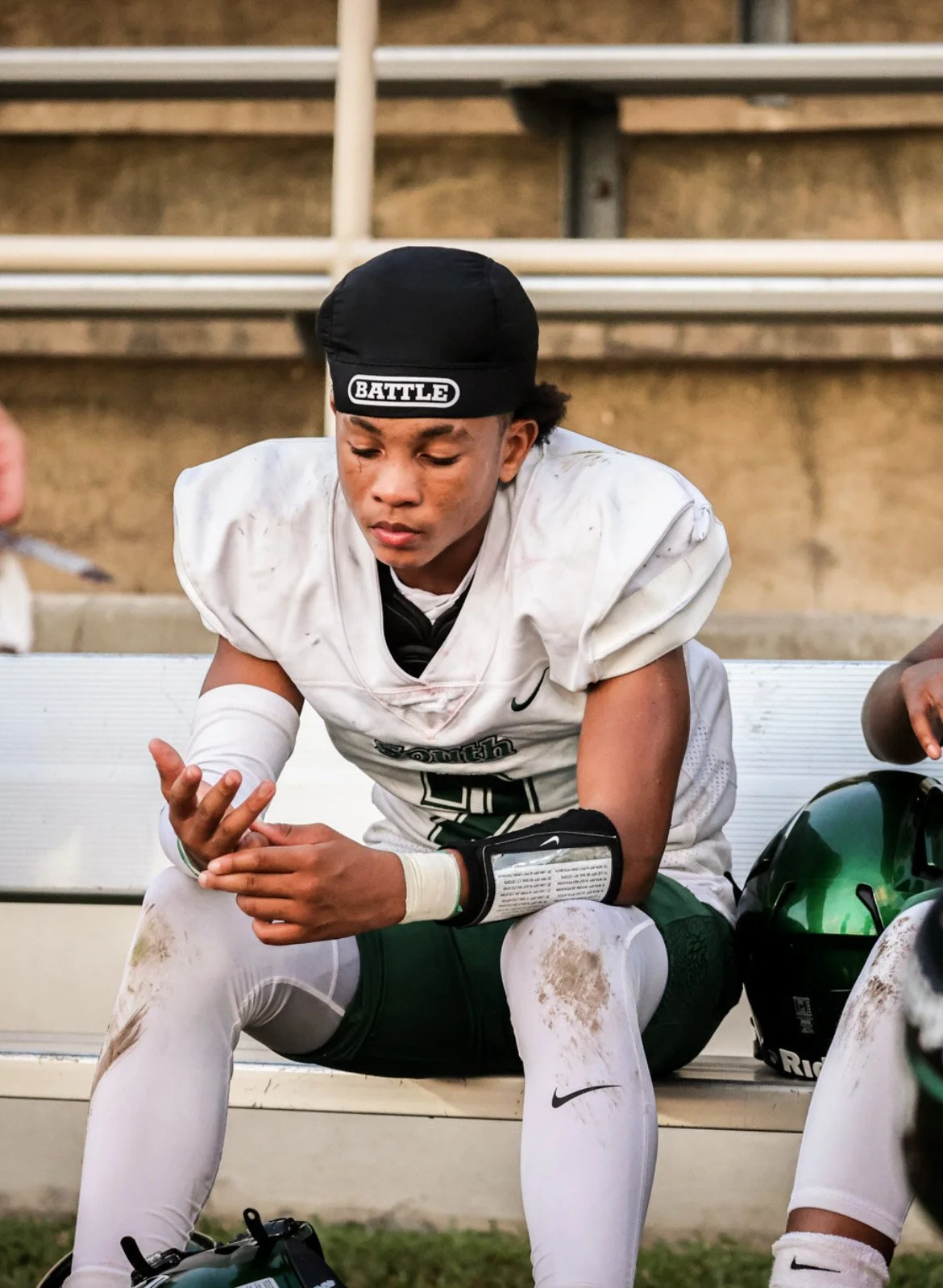 A young football player sits on a bench in uniform, adjusting his wristband, with a helmet beside him.