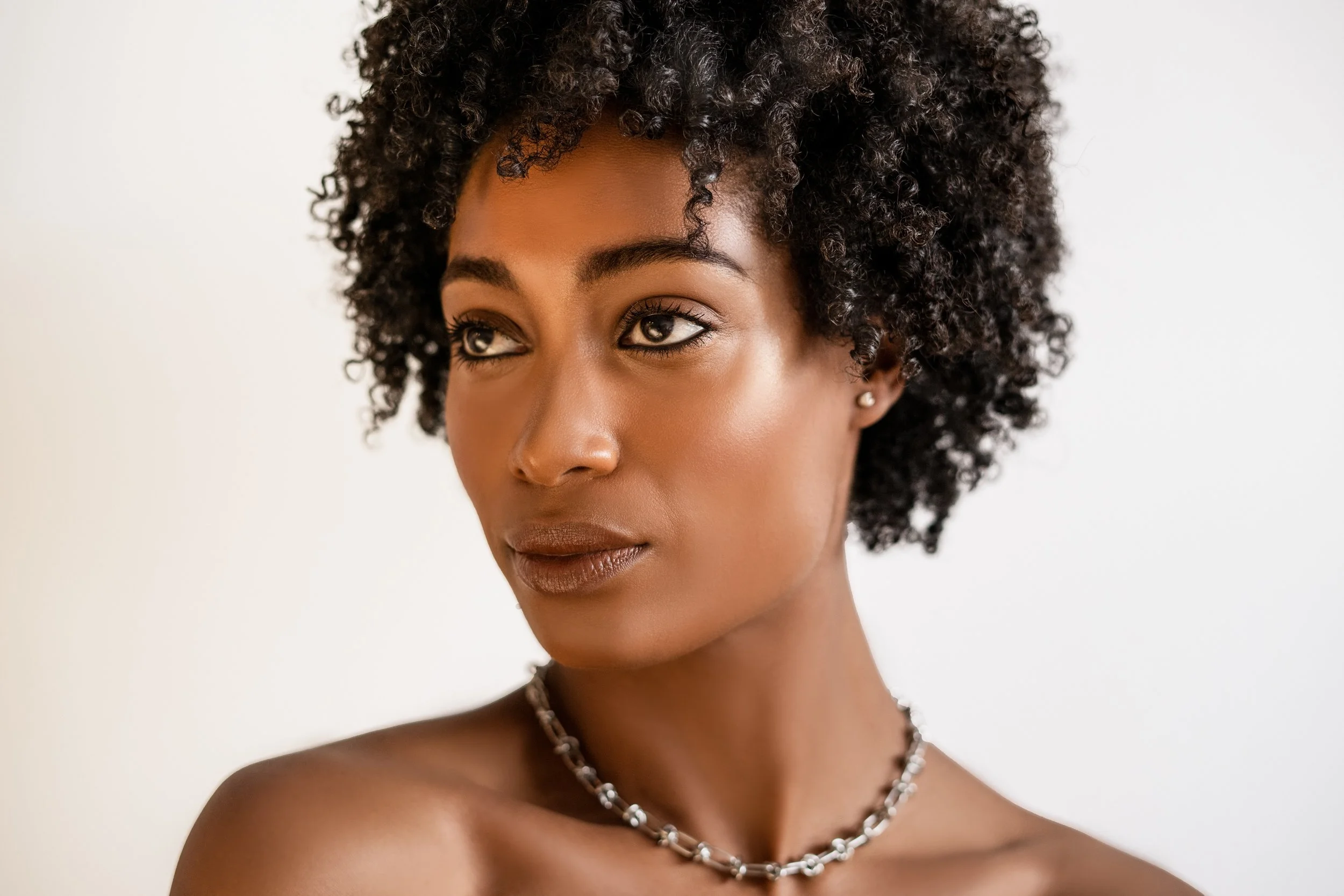 Close-up portrait of a woman with natural curly hair, wearing minimal makeup and a pearl necklace, looking slightly to the side against a plain white background.