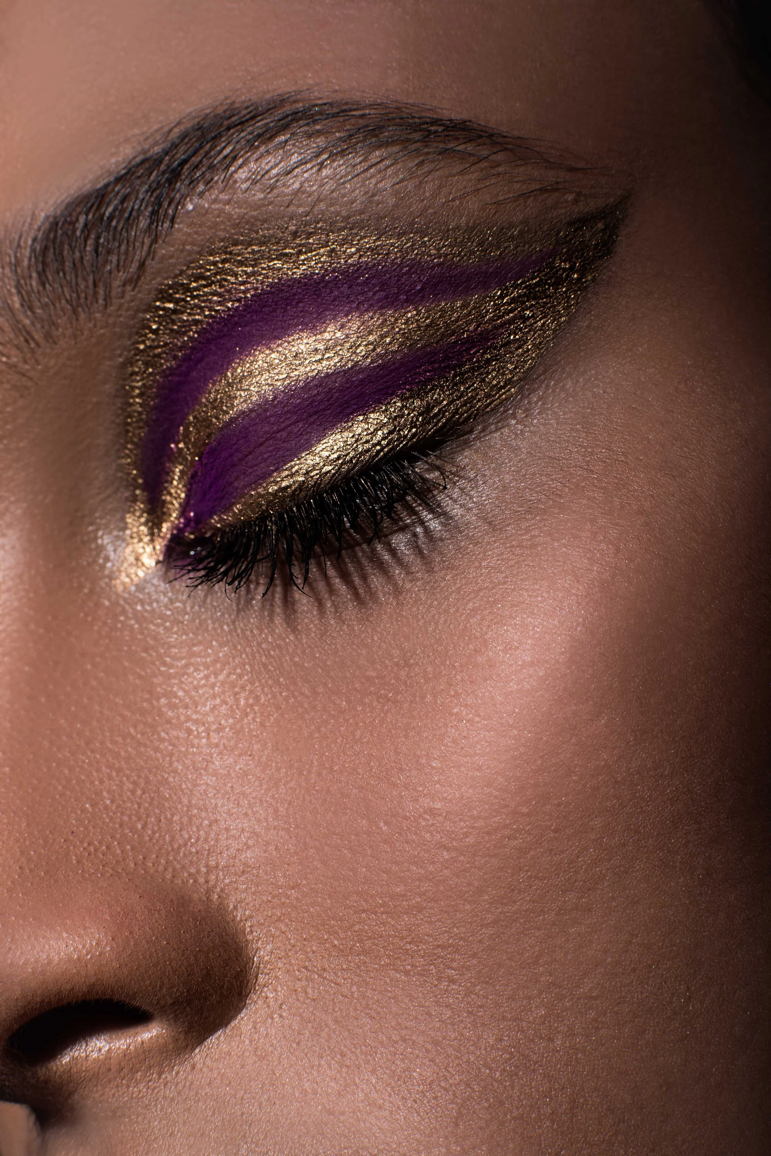 Close-up of a person's eye with dramatic eye makeup, featuring gold glitter eyeshadow, purple eyeliner, and black eyelashes.