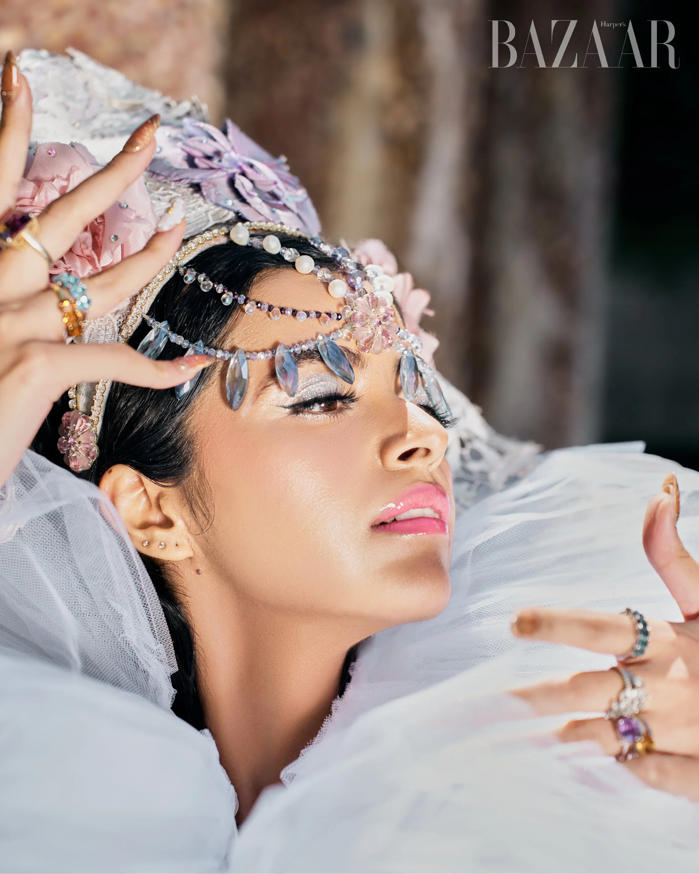 A woman wearing jewelry and a white tulle outfit, lying down with her head resting on a pillow, looking thoughtfully at the camera.