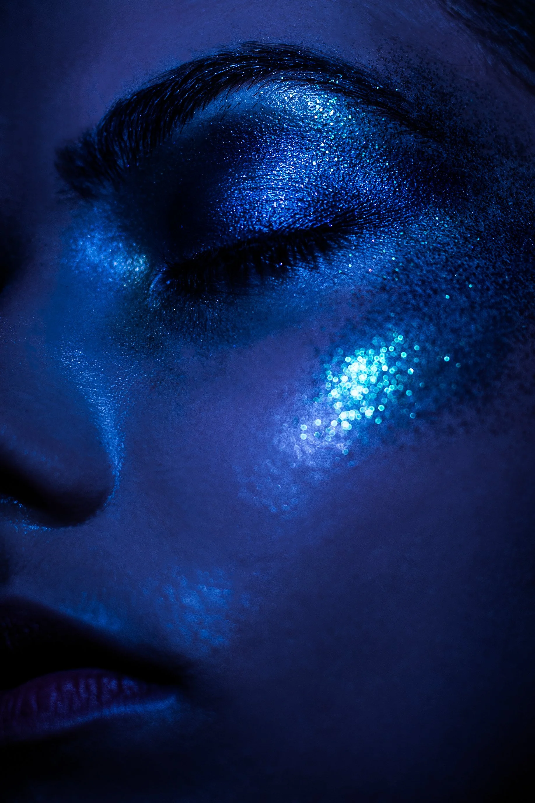 Close-up of a person's face with closed eye, covered in blue glitter makeup under blue lighting.