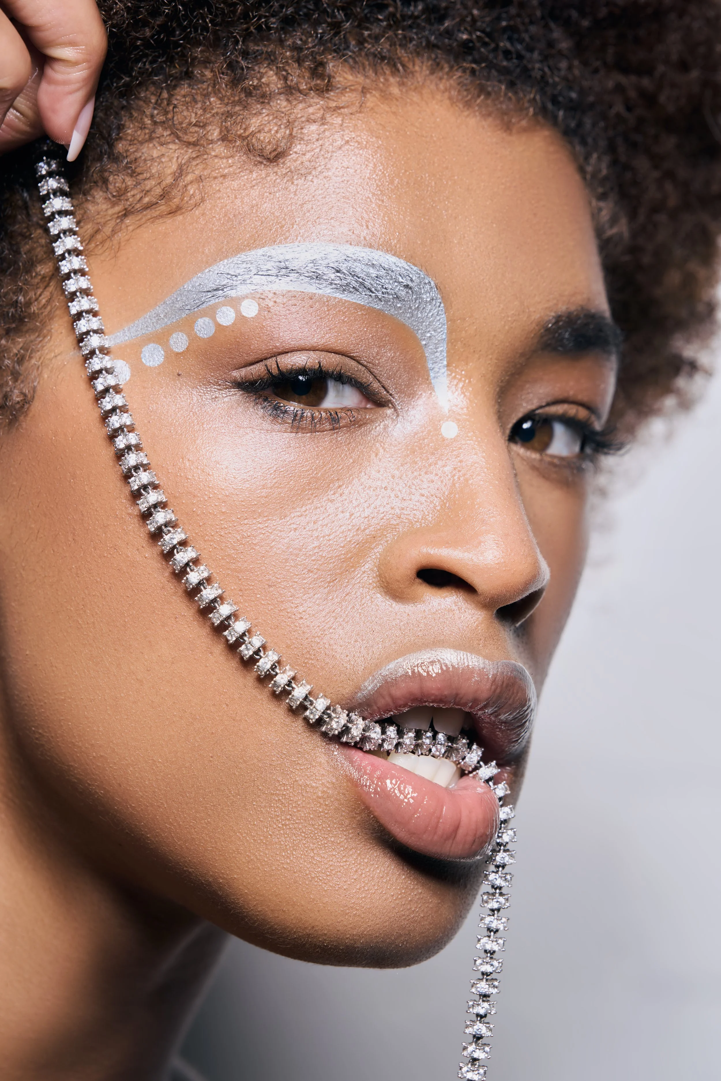 Close-up of a woman's face with artistic white makeup, holding a diamond necklace in her mouth, against a neutral background.