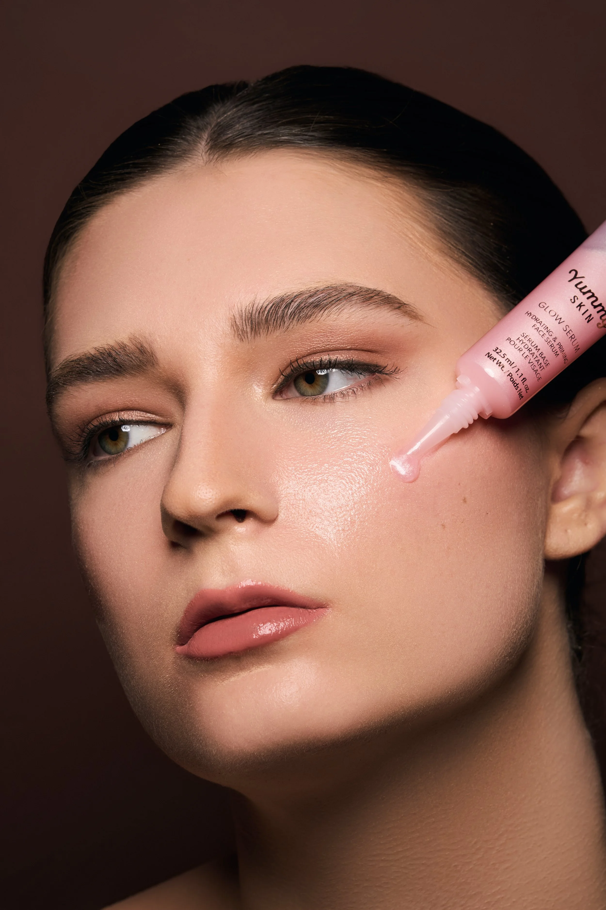 A woman applying a pink facial serum to her cheek, with clear skin and natural makeup, against a dark background.