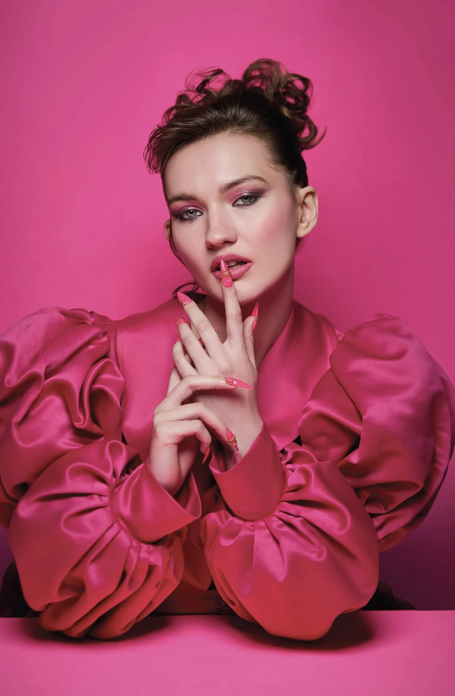 A woman with styled hair and pink makeup poses against a pink background, wearing a pink satin blouse with puffy sleeves, and has matching pink artfully decorated nails, touching her lips with one finger.