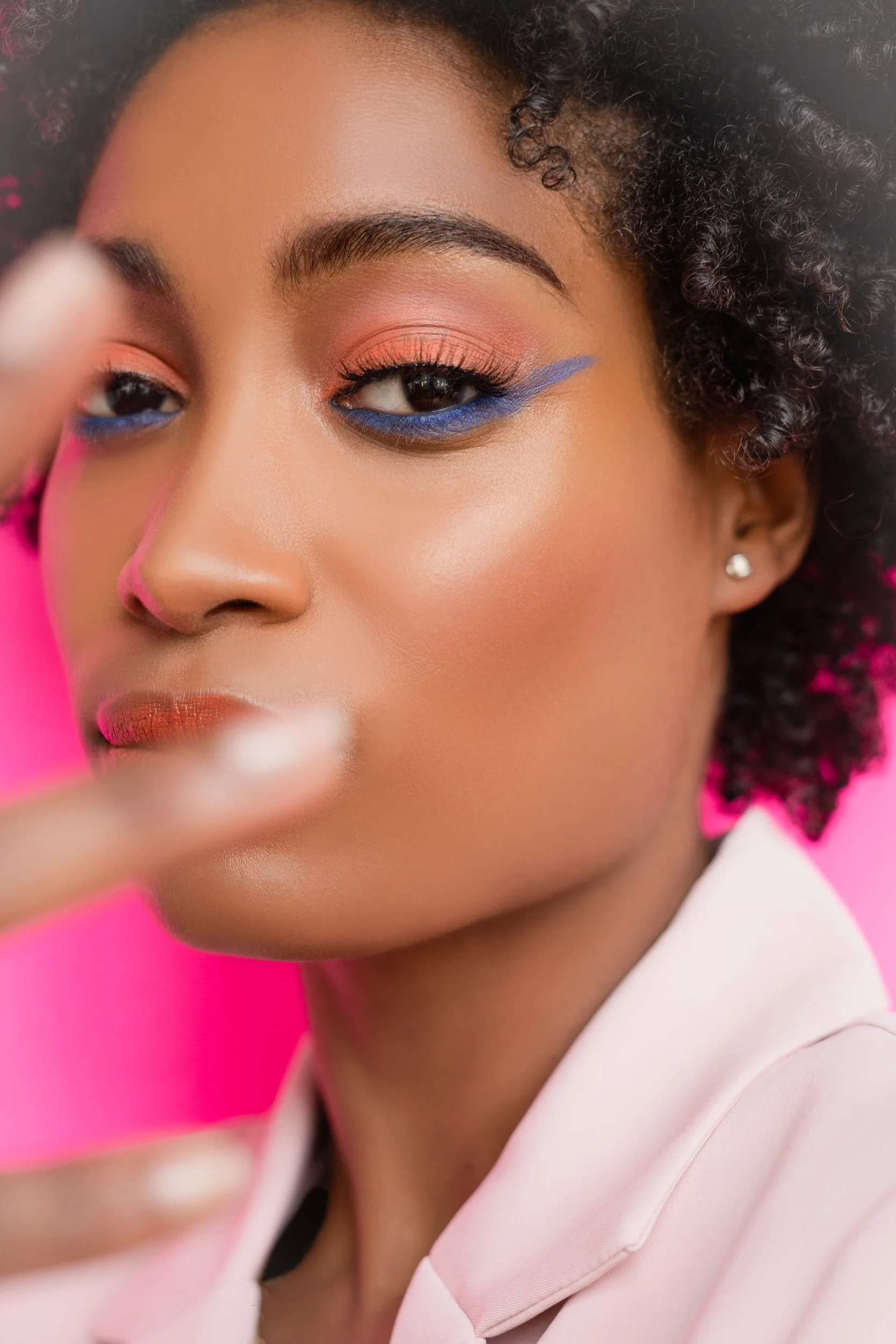 Close-up of a woman with curly hair, colorful makeup, and a soft pink background.