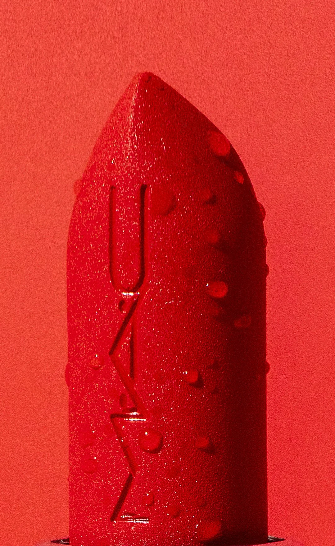 Close-up of a red lipstick with water droplets on its surface, set against a red background.