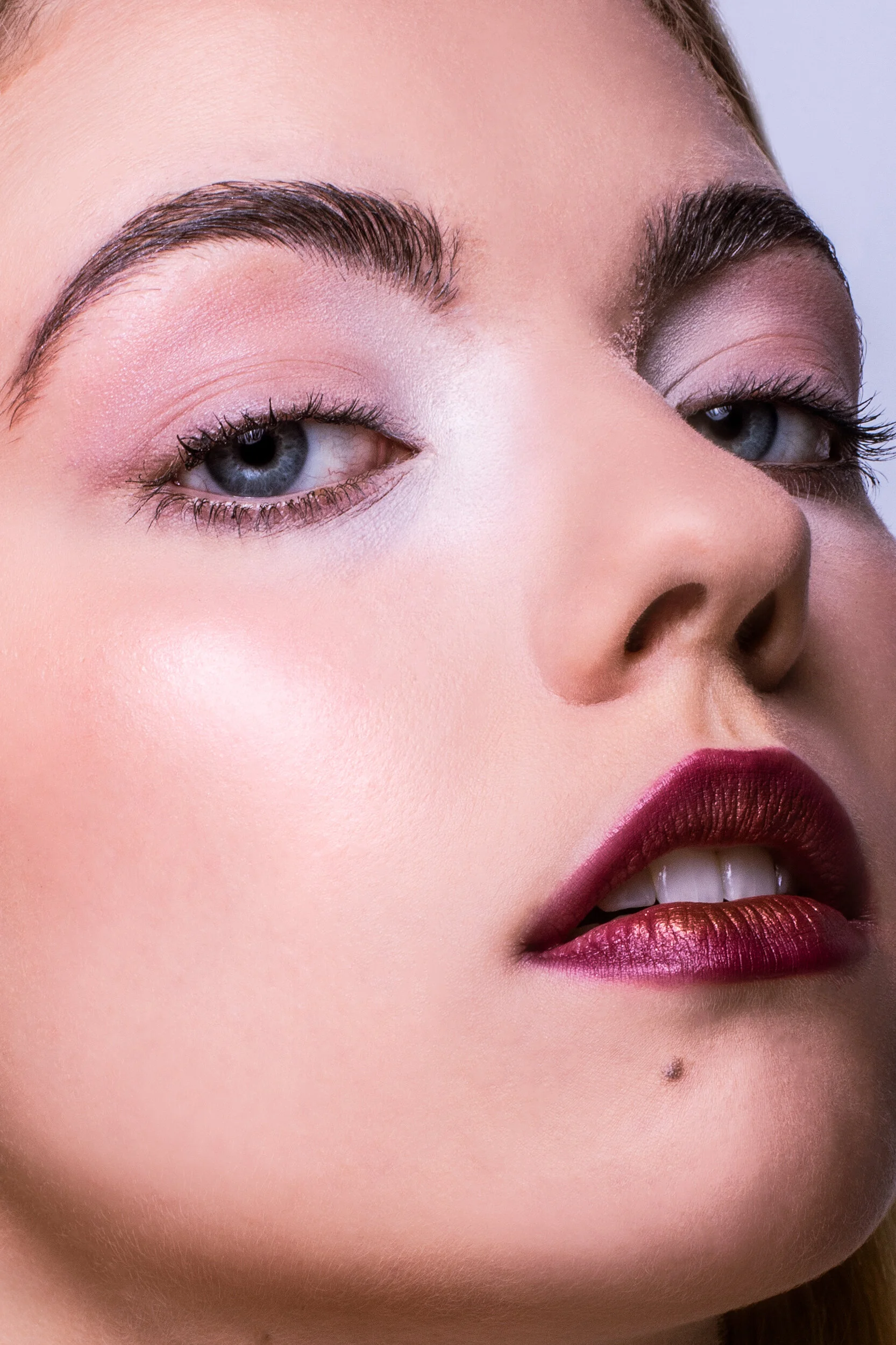 Close-up of a woman's face focusing on her eye, cheek, nose, and lips, with makeup and a beauty mark.