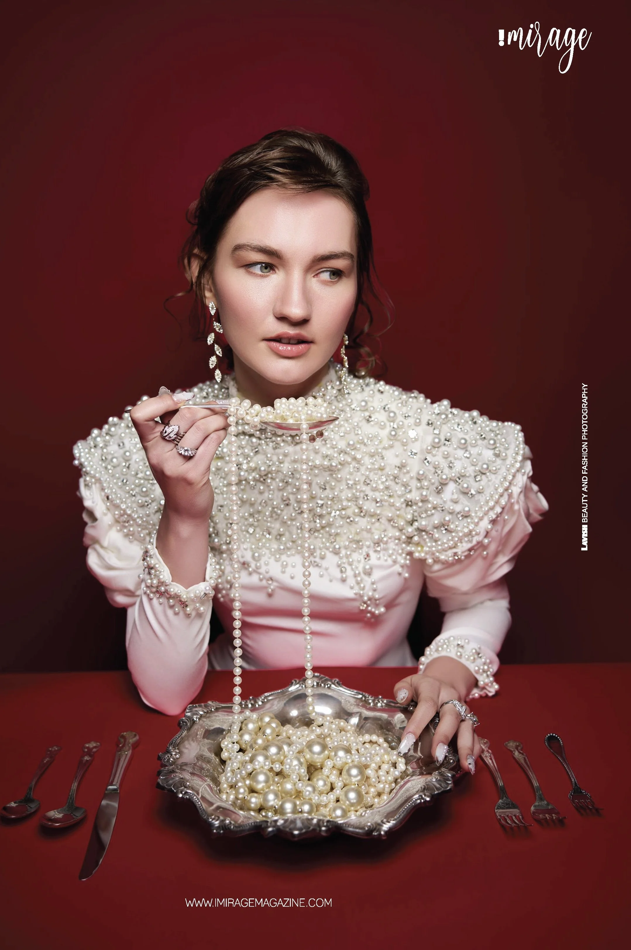 A woman dressed in an elegant, pearl-embellished white gown sitting at a table with a silver bowl filled with pearls. She is holding a spoon with pearls, wearing jewelry, and has a red background.
