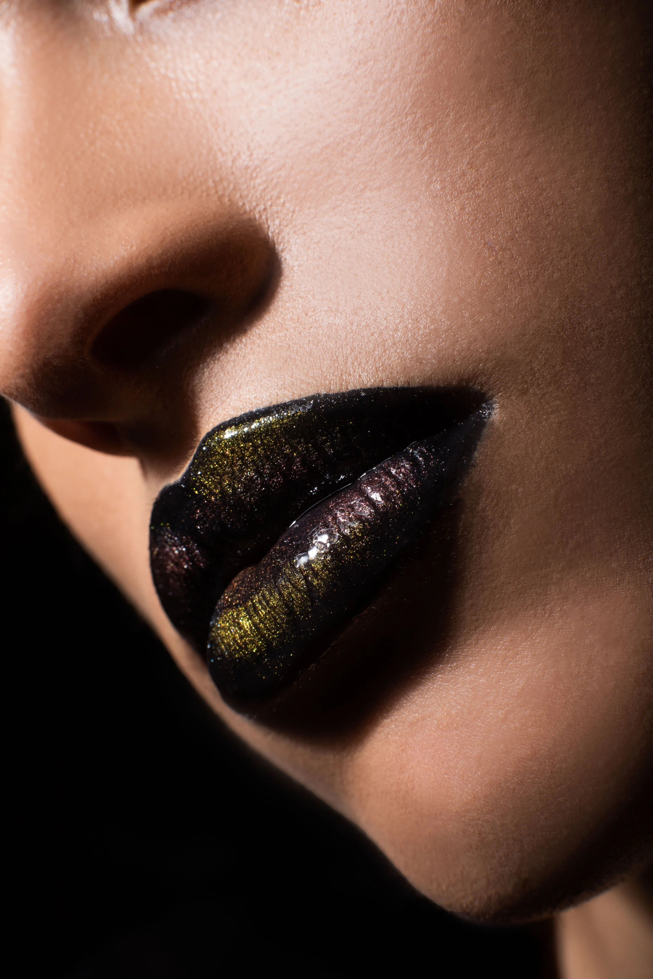Close-up of a person's lips with black lipstick with gold shimmer, part of their nose and cheek are visible.