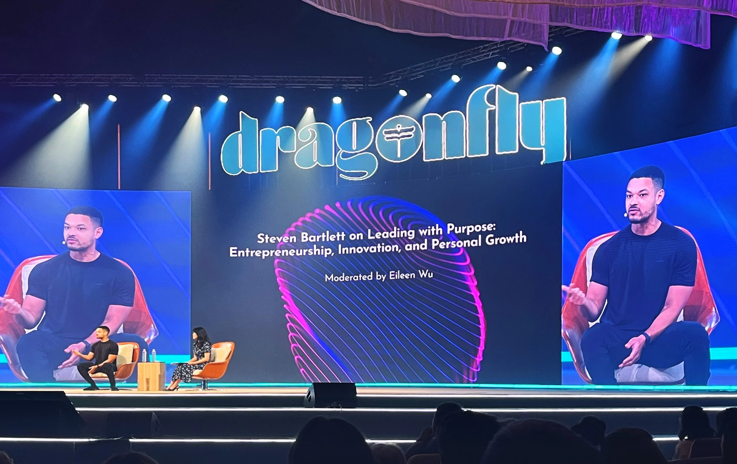 Stage at conference with a large screen displaying 'dragonf' and a speaker, Steven Bartlett, seated, with text about leadership, entrepreneurship, innovation, and personal growth. Two people seated on stage, one speaking, and a third person standing.