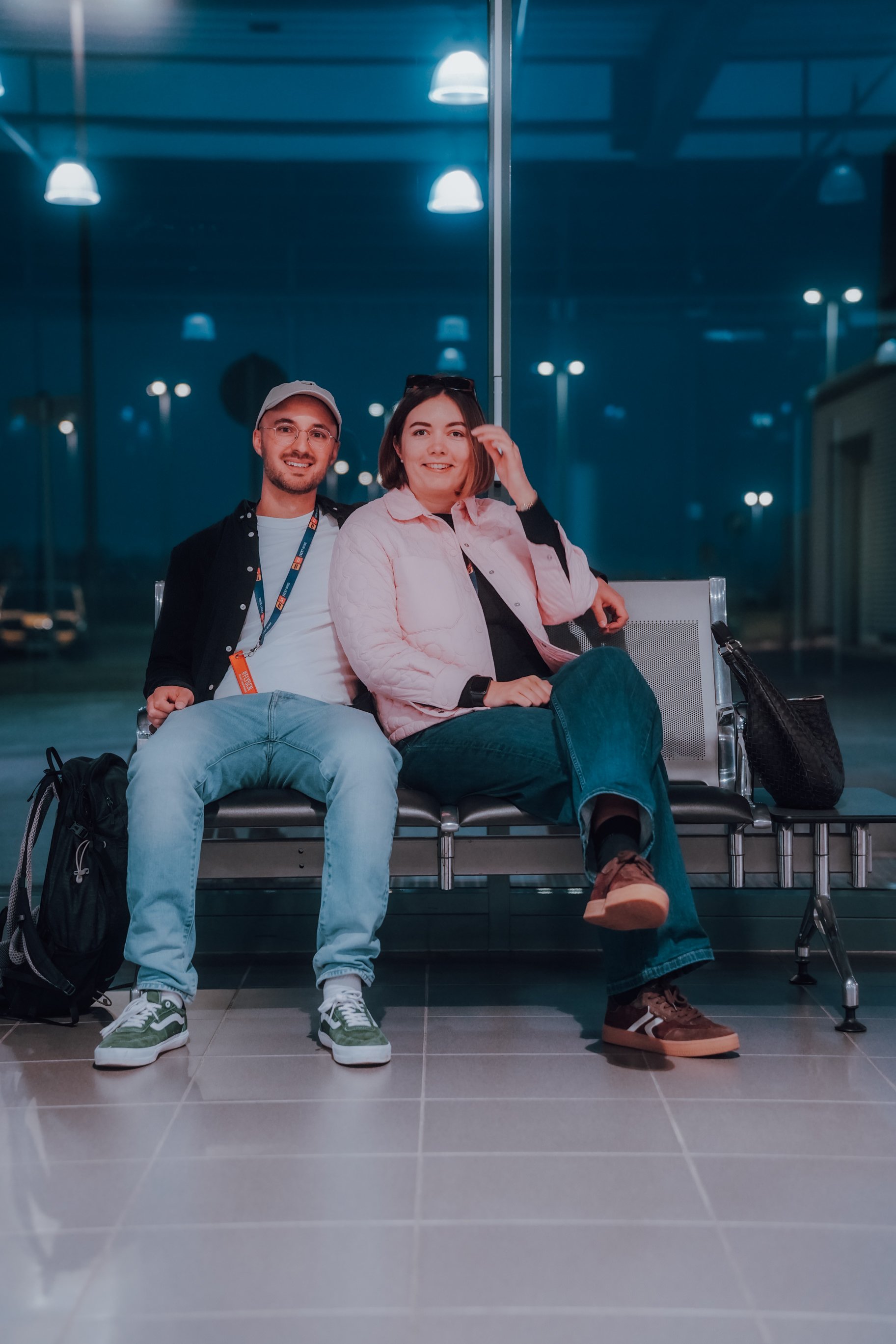 Marcel und Victoria sitzen im Flughafen. Es ist Nacht, und sie haben Koffer und Rucksäcke bei sich. Hinter ihnen große Glasfenster, reflektierende Deckenleuchten und eine ruhige Atmosphäre.