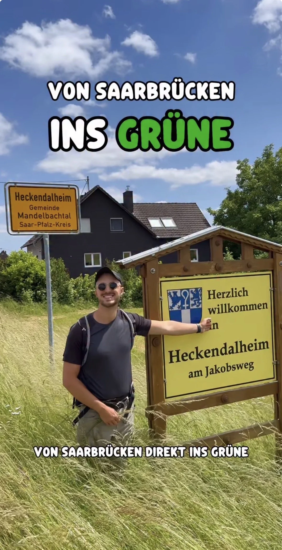 Ein junger Mann mit Sonnenbrille und Rucksack steht neben einem Hinweisschild in einem grünen Feld. Über dem Bild steht in großen Buchstaben: 'Von Saarbrücken ins Grüne'. Ein gelbes Ortsschild zeigt 'Heckendalheim, Gemeinde Mandelbachtal, Saar-Pfalz-Kreis', und das Holzschild daneben begrüßt Besucher mit 'Herzlich willkommen in Heckendalheim am Jakobsweg'.