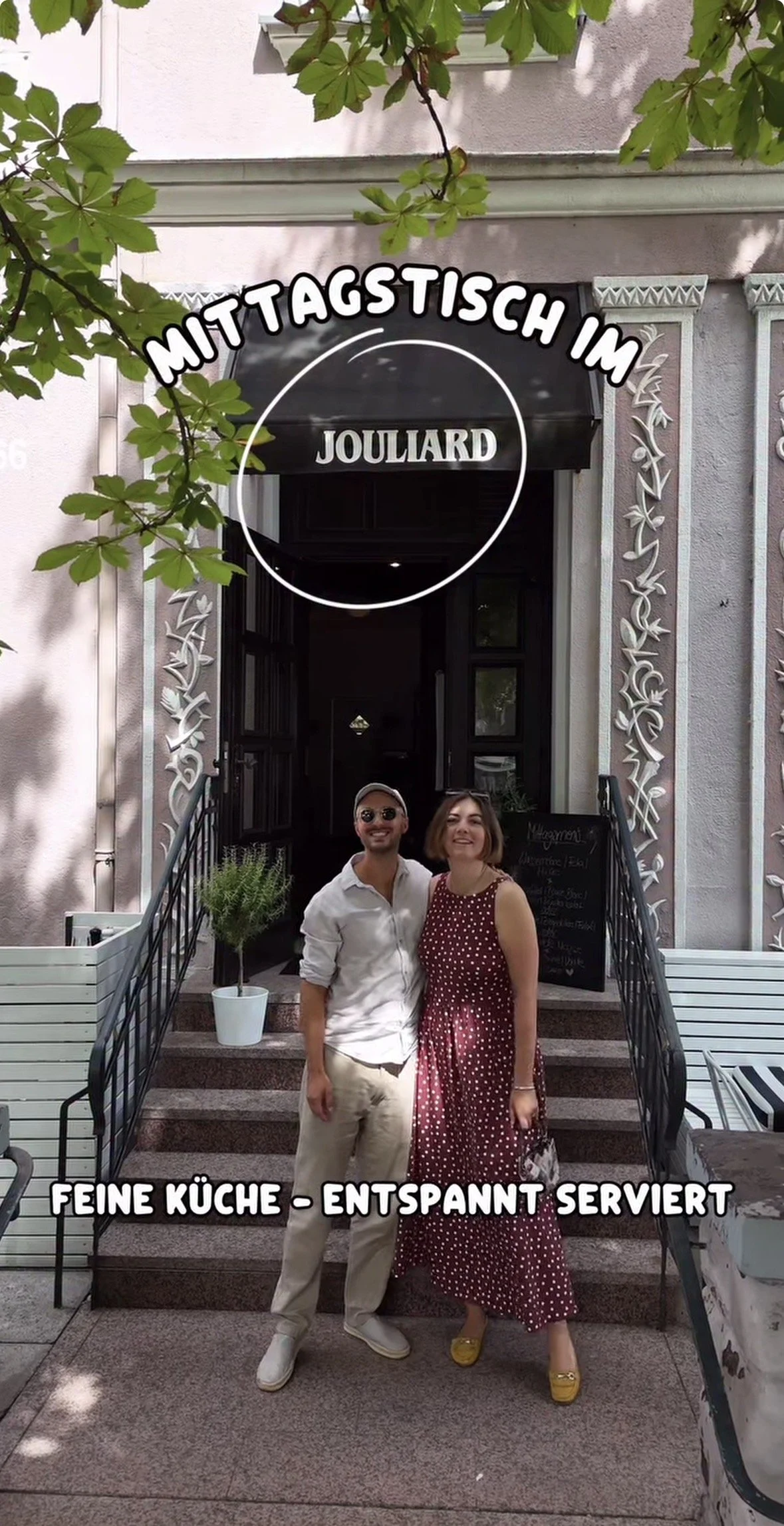 Zwei Menschen, ein Mann und eine Frau, stehen auf den Stufen vor einem Restaurant namens "Joullard". Es ist tagsüber, und um sie herum sind Bänke, Pflanzen und dekorative Fassaden. Über ihnen ist ein Baum mit grünen Blättern, die Schatten werfen.