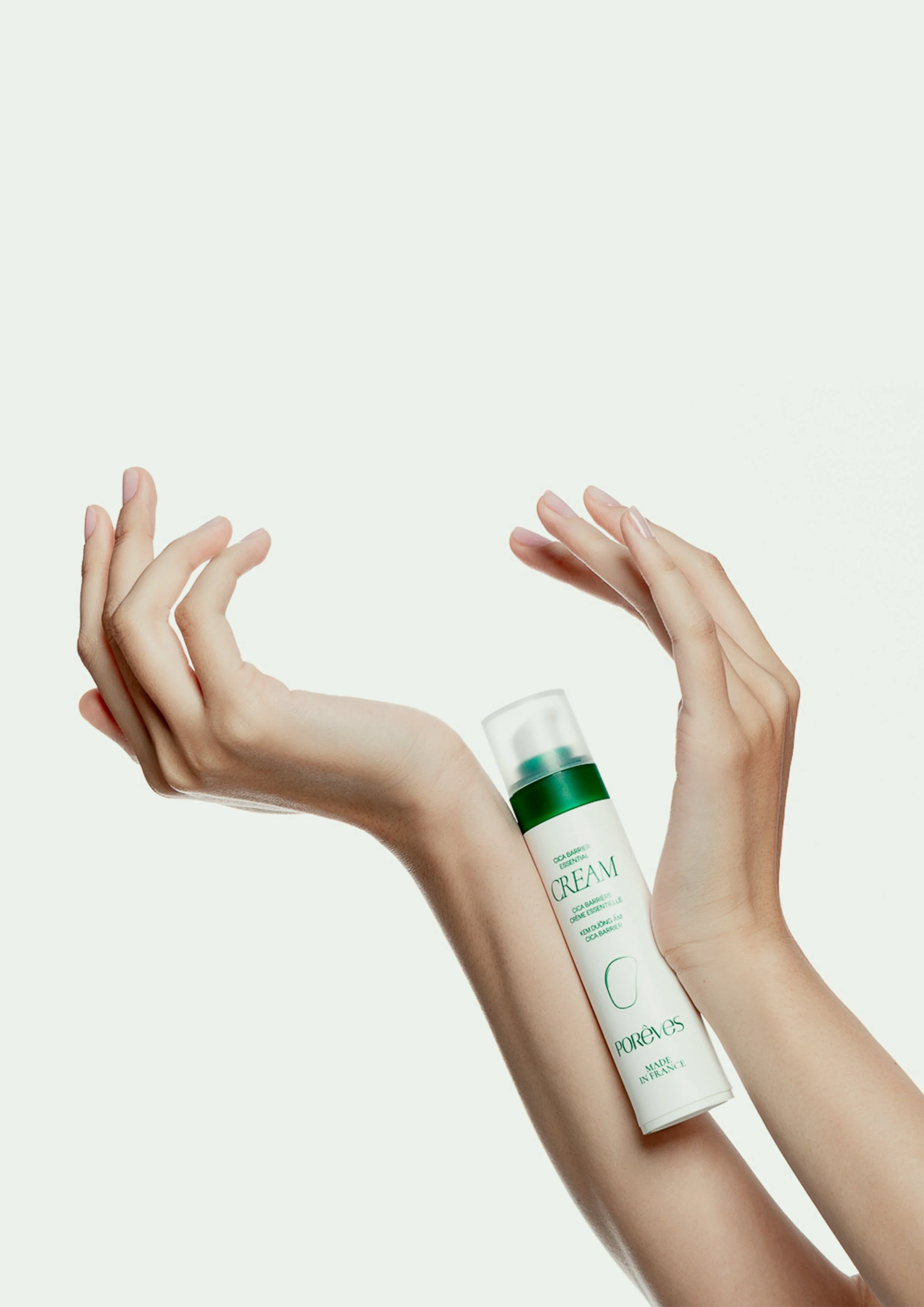 Des mains d’une personne à la peau claire : l’une tenant un flacon de crème blanc avec des accents verts de la marque PORÊVES designé par le studio impulso, l’autre délicatement courbée, sur un fond clair et uni.