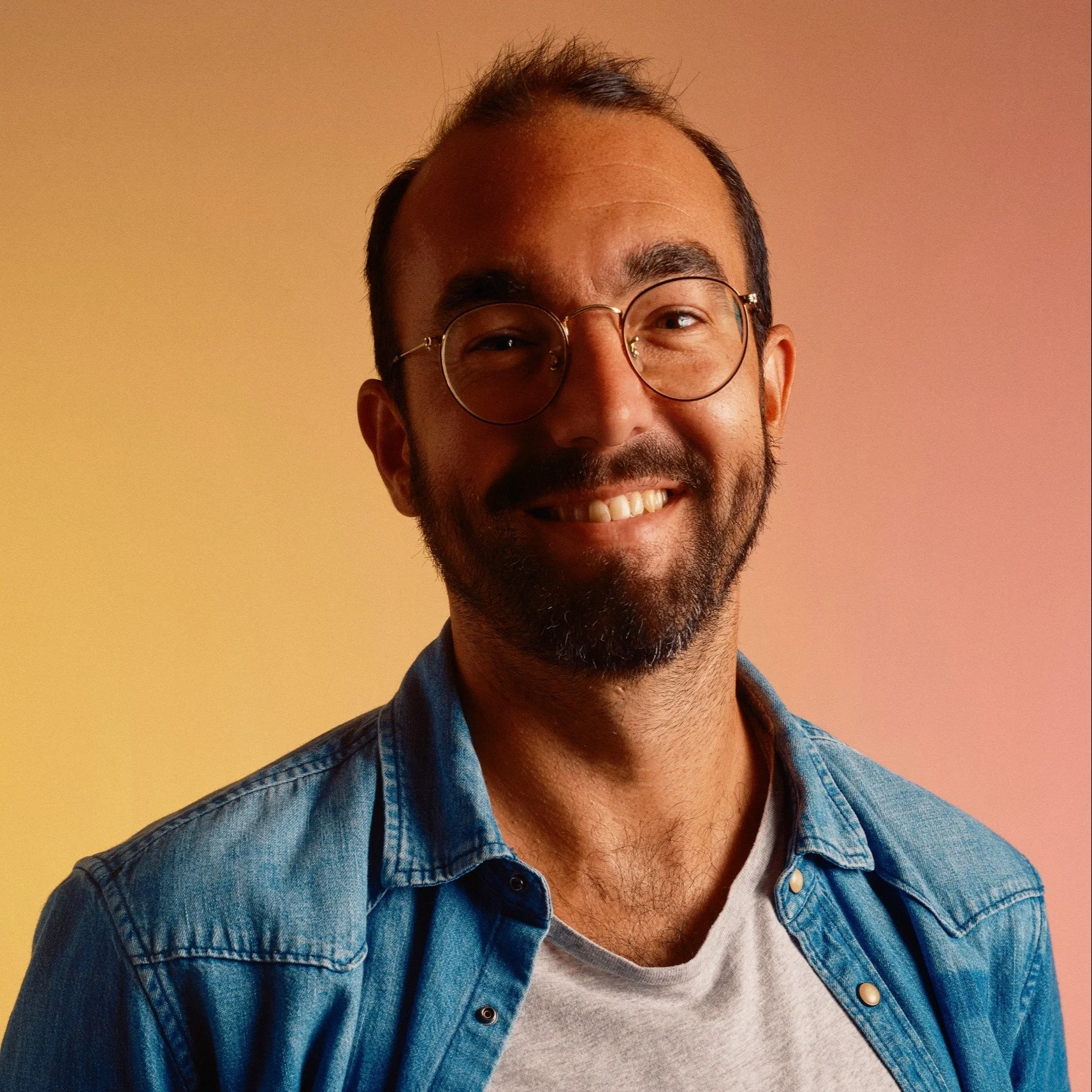 Smiling man with glasses, beard, and mustache wearing a denim shirt over a light-colored T-shirt, against a gradient background of warm tones.