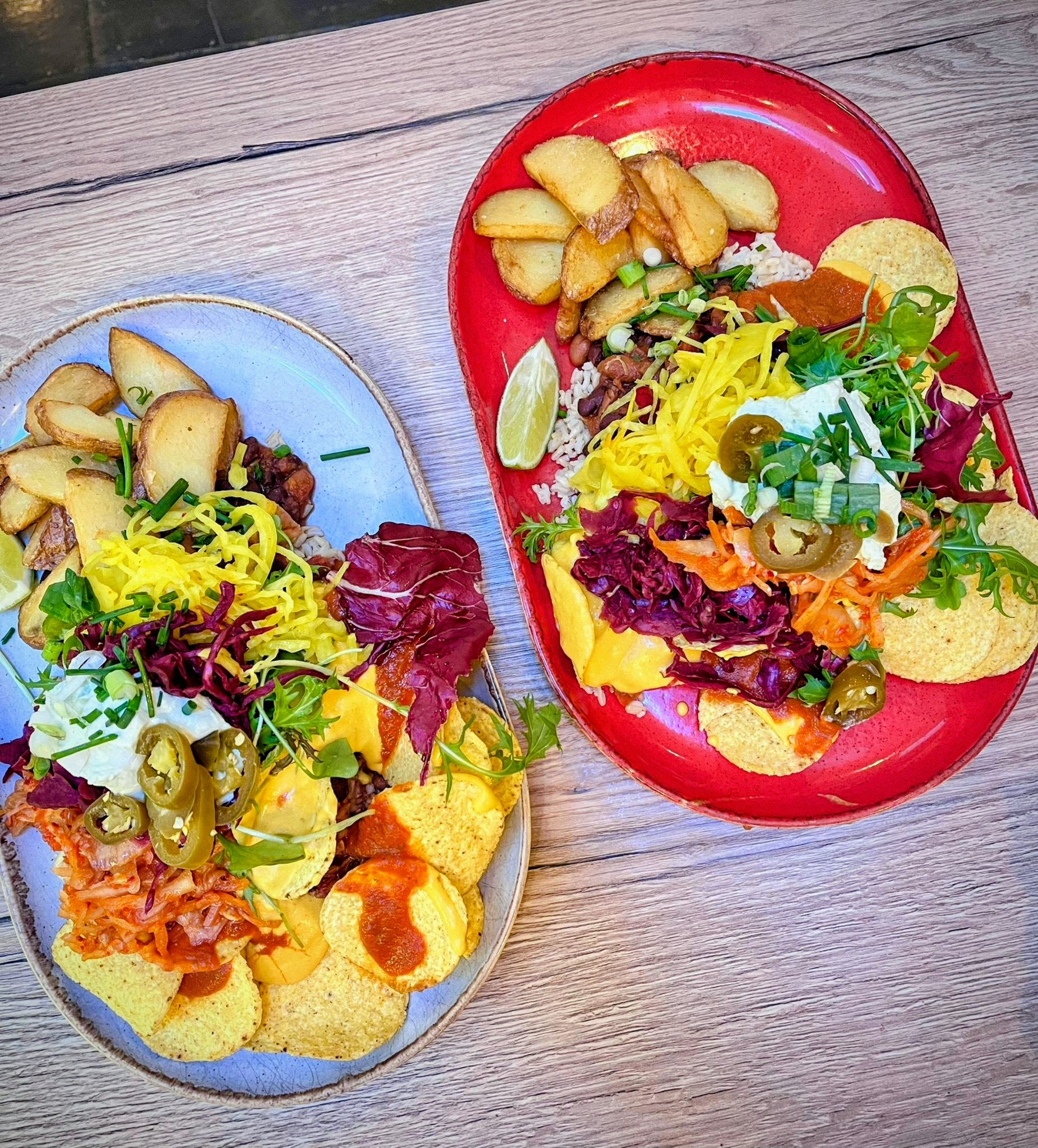 Two colorful plates of our award winning nachos with various toppings including nacho chz sauce, seasonal leaves, curtido, jalapenos and sour cream, alongside potato wedges, on a wooden table.