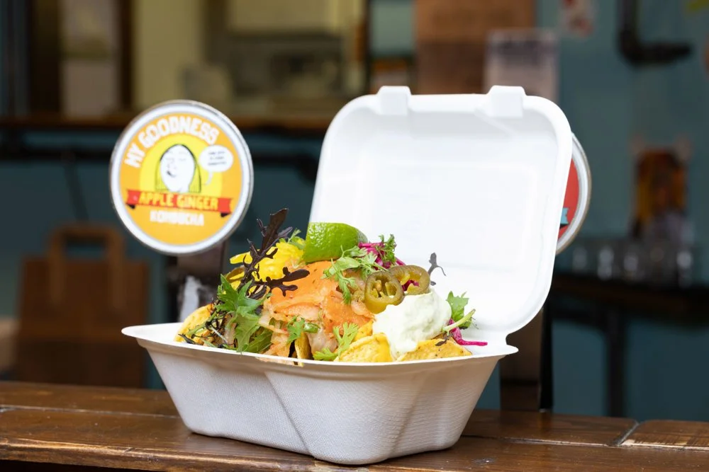 A takeout container filled with our award winning vegan nachos, garnished with lime, jalapeños, and leafy greens, sitting on a wooden surface at the English Market stall.