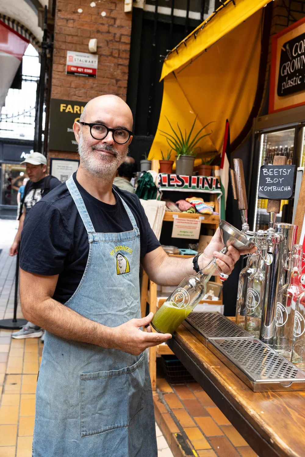 A man with glasses and a gray beard wearing a black T-shirt and light blue apron standing at a beverage stand, pouring some kombucha in to a glass My Goodness bottle at the English Market.