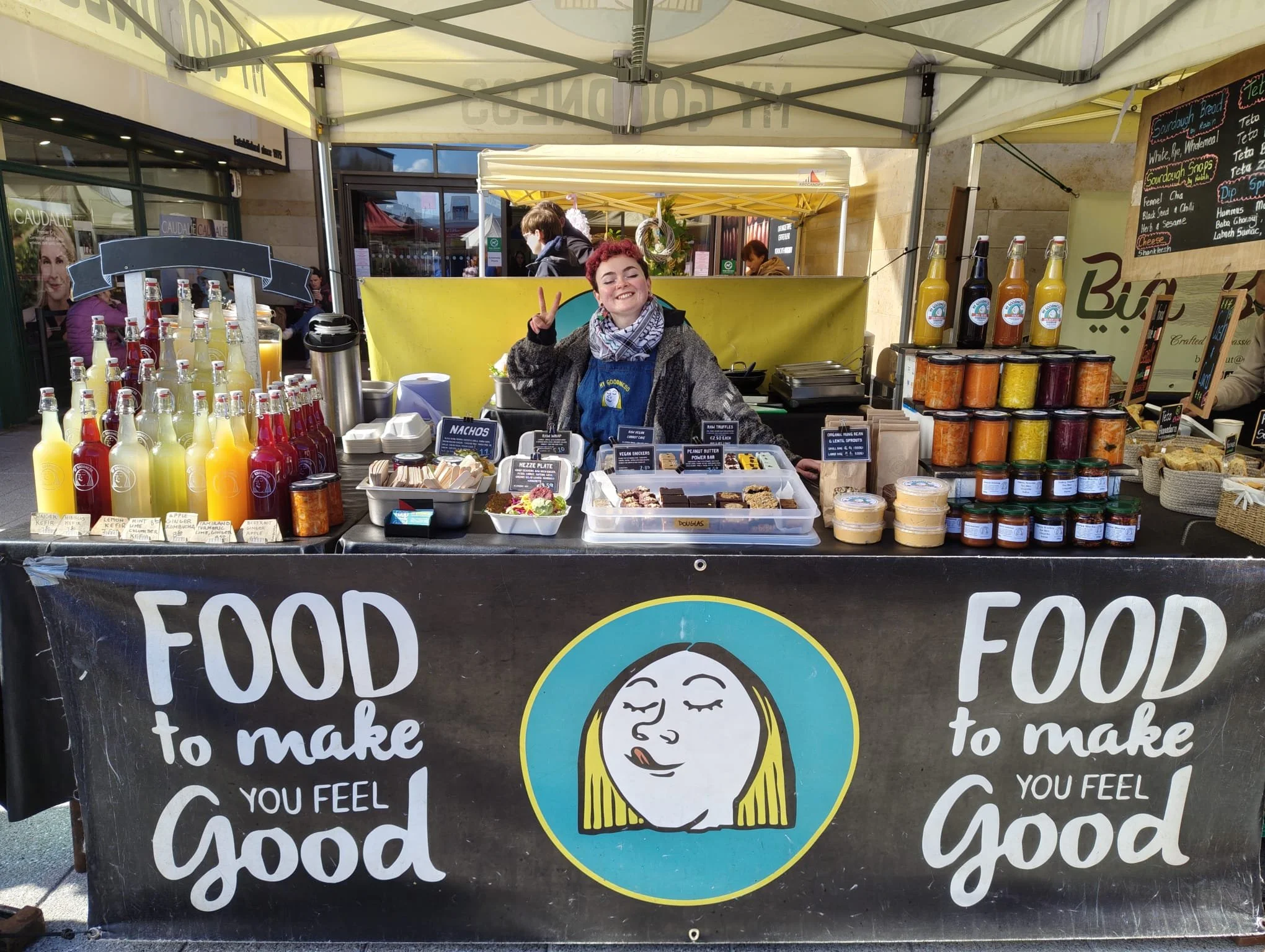 Smiling woman with short red hair making a peace sign at the My Goodness farmers market stall.