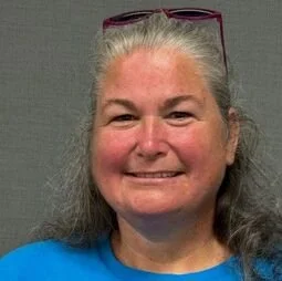 Close-up of a smiling woman with gray hair wearing sunglasses on her head and a blue shirt.