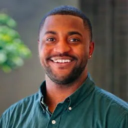 A smiling man with short hair and a beard, wearing a green button-down shirt, standing outdoors with a blurred background of trees and a building.