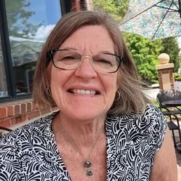 A smiling middle-aged woman with glasses, short brown hair, wearing a patterned top, sitting outdoors with an umbrella and greenery in the background.