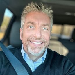 A middle-aged man with gray hair and a beard smiling, sitting in a car, wearing a blue shirt and a dark sweater.