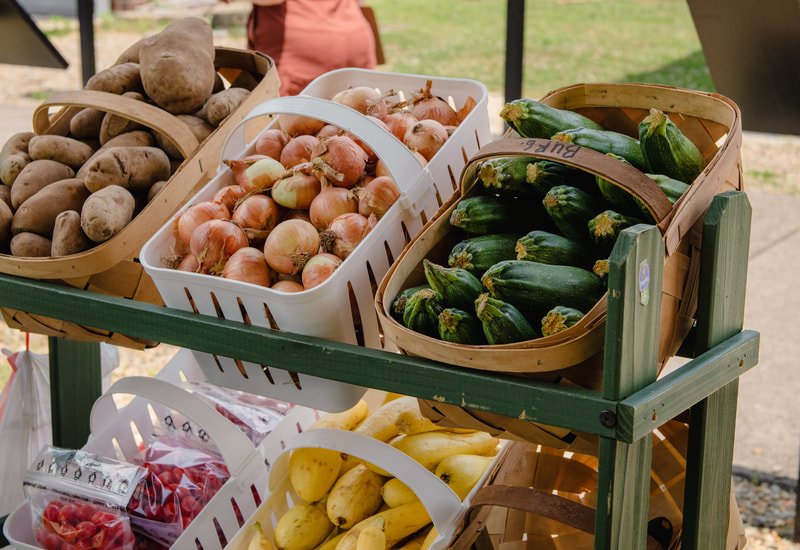 Fresh vegetables including potatoes, onions, zucchinis, and bananas displayed on a green-finished wooden stand at a farmers market.